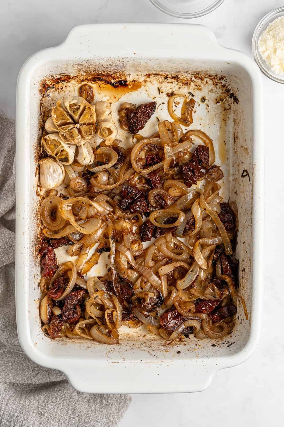 A white baking dish filled with roasted onion slices, garlic cloves, and sun-dried tomatoes. The ingredients are browned, with areas of char on the edges. A gray cloth is partially visible on the left side.