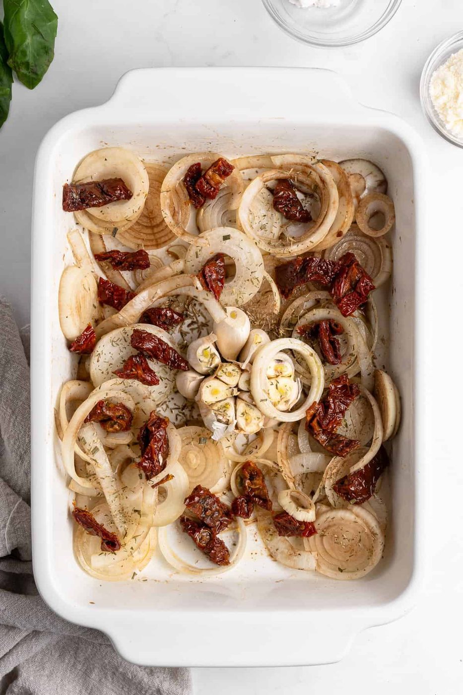 A white baking dish filled with layered onion slices, sun-dried tomatoes, and a halved bulb of garlic, all seasoned with herbs. The dish is ready for baking, surrounded by a beige cloth and small bowls of ingredients on a white marble surface.