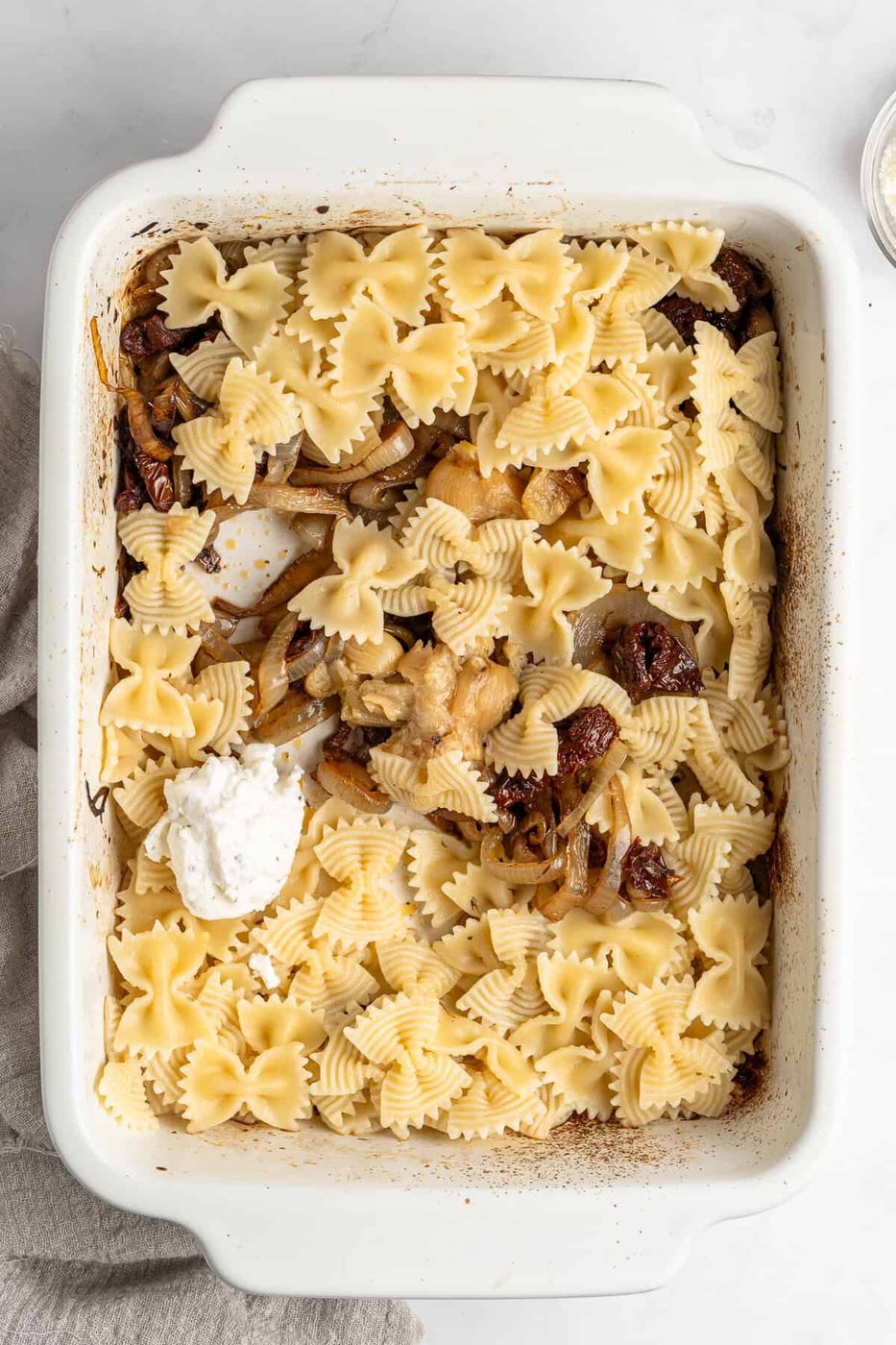 A white baking dish filled with bowtie pasta, sautéed onions, sun-dried tomatoes, and a dollop of ricotta cheese on top. The edges of the dish show slight browning, indicating baking. A beige cloth is partially visible on the side.