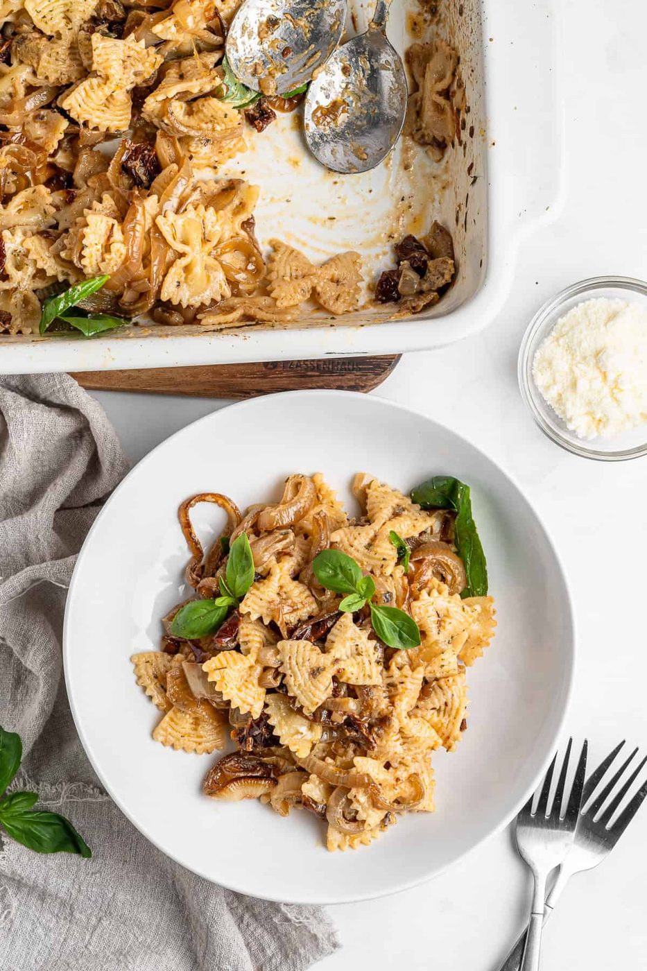A white plate with bow-tie pasta mixed with caramelized onions, mushrooms, and sundried tomatoes, garnished with fresh basil. Next to it is a baking dish with more pasta, two spoons, a napkin, grated cheese in a bowl, and two forks.