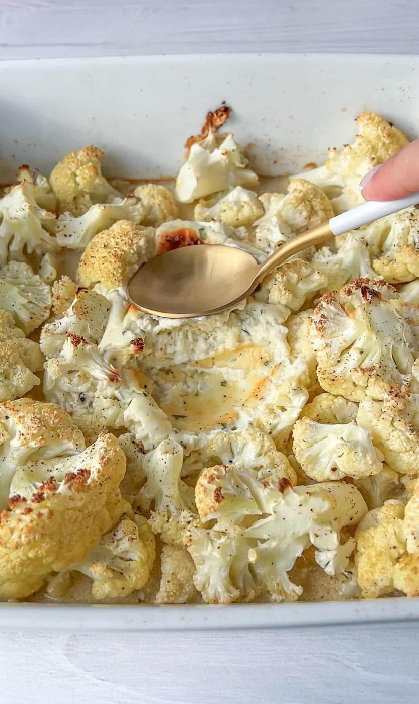 A hand holds a spoon, scooping creamy cheese sauce over roasted cauliflower florets in a white baking dish. The cauliflower is lightly browned and seasoned with herbs, creating a savory dish.