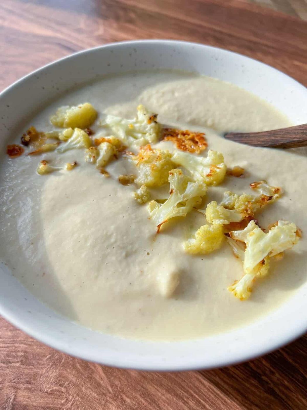 A bowl of creamy cauliflower soup garnished with roasted cauliflower florets. A wooden spoon rests in the bowl. The soup has a smooth texture, and the dish is placed on a wooden table surface.