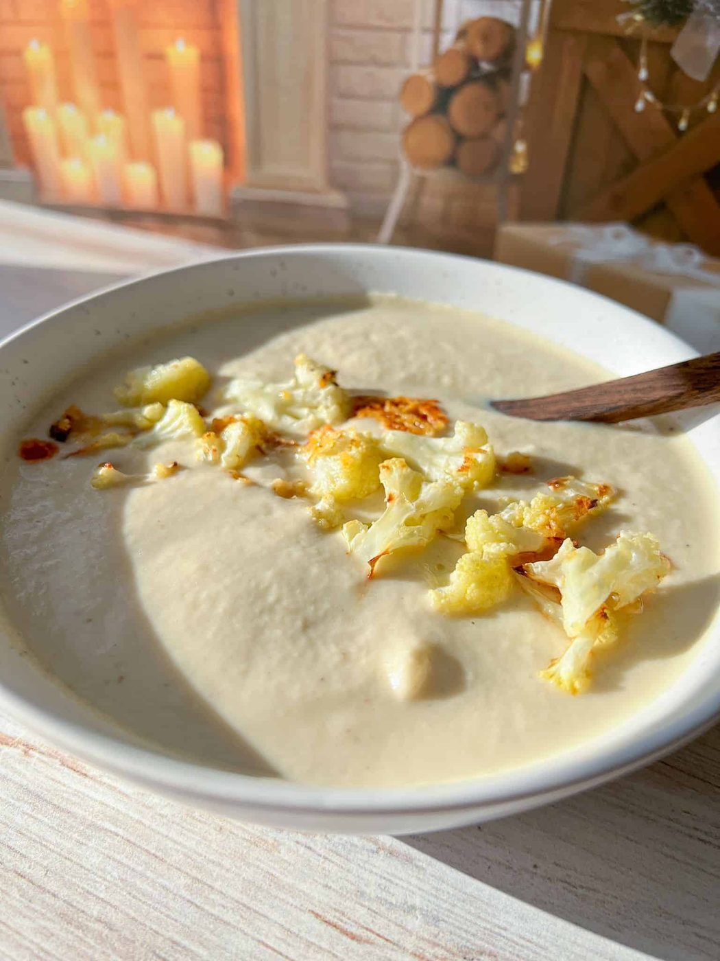 A bowl of creamy cauliflower soup topped with roasted cauliflower florets. A wooden spoon rests in the bowl. In the background, there's a cozy setting with candles and stacked firewood.