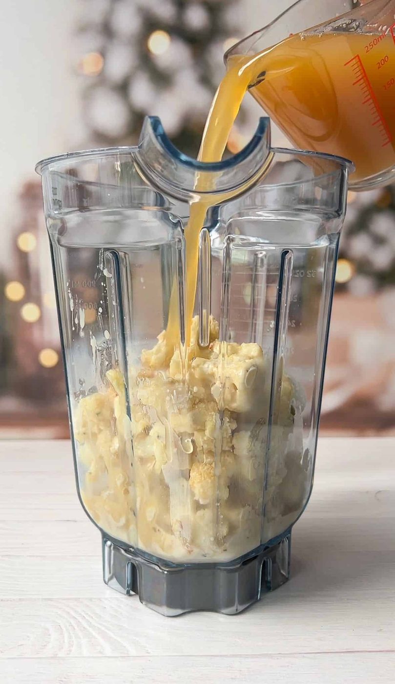 A clear blender jar filled with cooked pasta is placed on a white surface. Broth is being poured into the jar from a measuring cup. A blurred background shows warm holiday lights and a decorated tree.