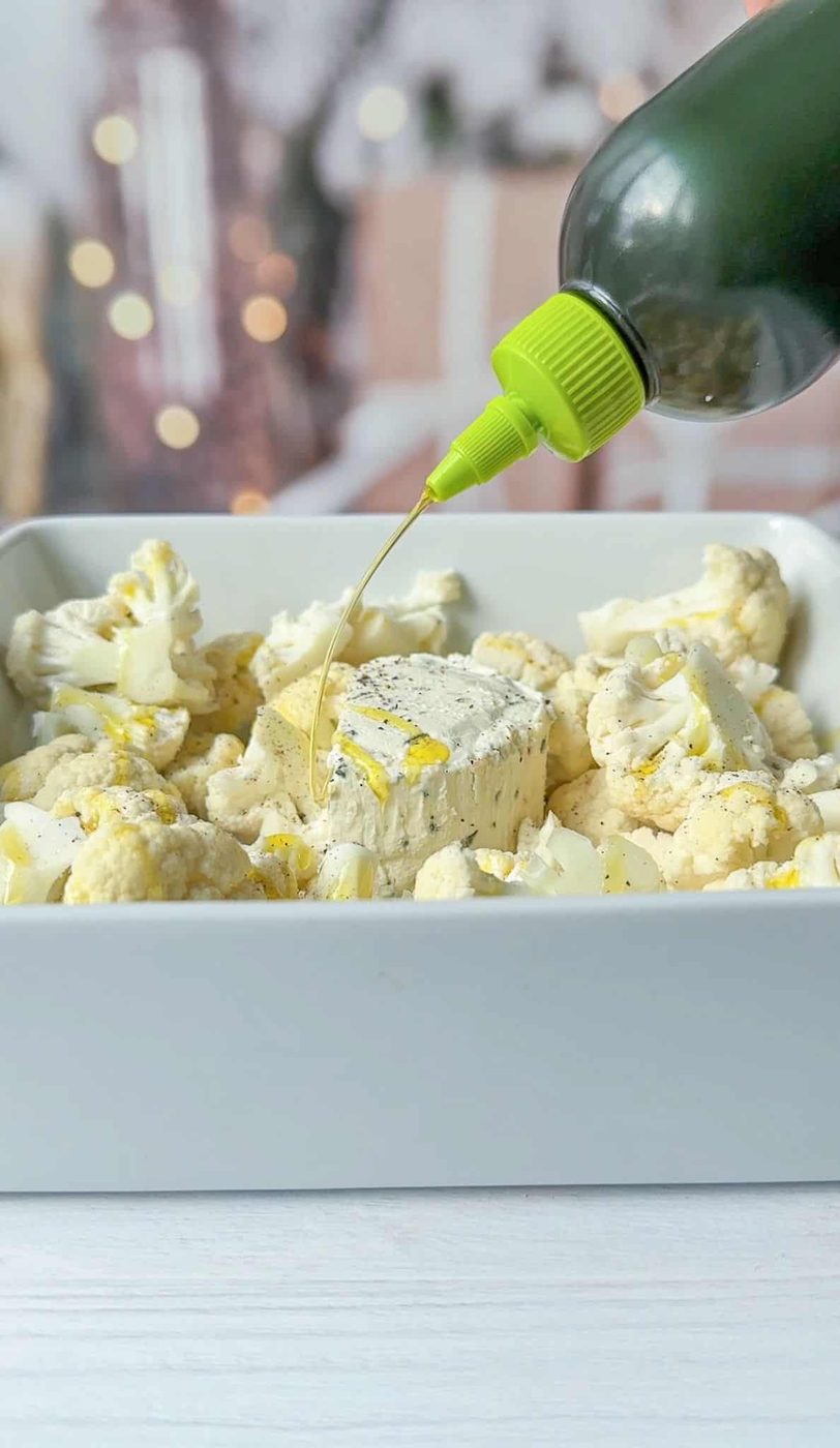 A bottle of olive oil is being drizzled over a square block of feta cheese, in a white baking dish. The background is softly blurred with hints of warm lights.