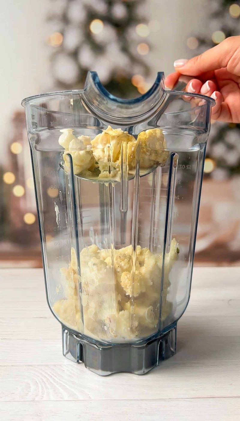 A blender jug with cauliflower pieces inside, held by a person. Blurry background with festive lights and trees.