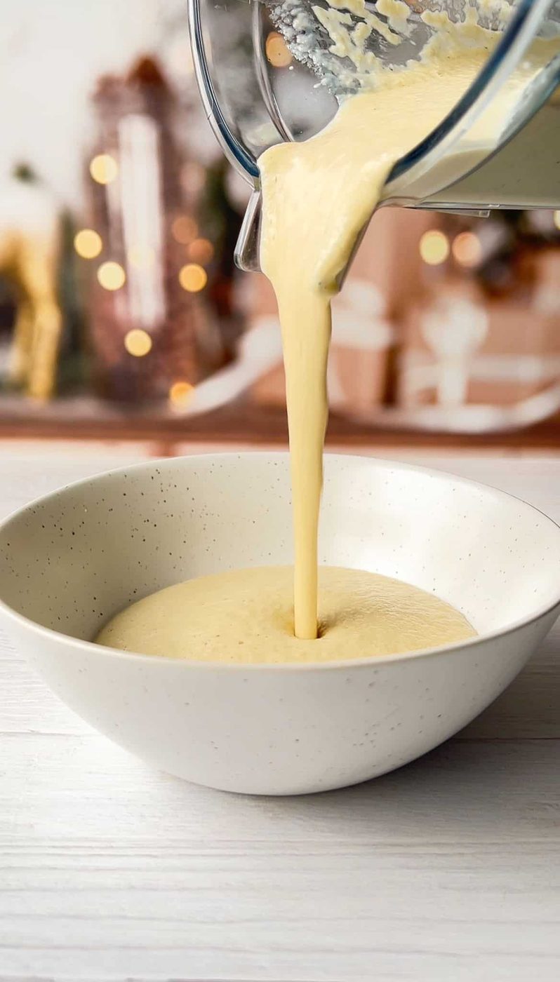 Pouring creamy yellow liquid from a blender jar into a speckled white bowl. The background is softly blurred, featuring warm lights and decorative elements. The scene suggests preparation for a recipe or meal.