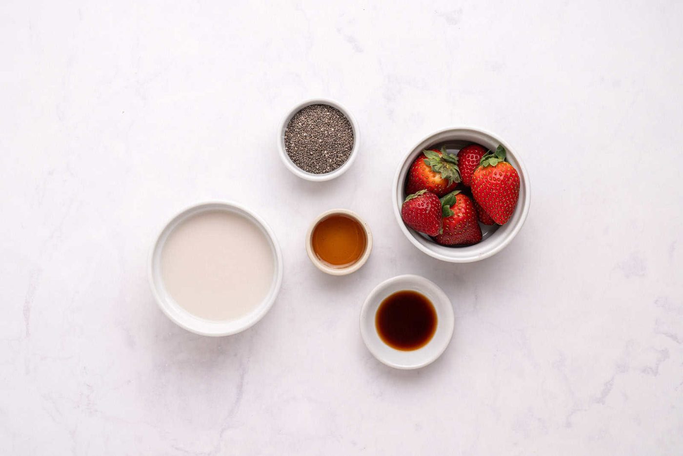 Five bowls on a white surface containing chia seeds, whole strawberries, plant-based milk, honey, and vanilla extract, arranged in a loose circle.