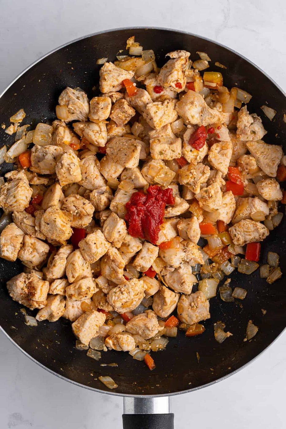 A saute pan with golden pieces of cooked, cubed chicken, as well as various spices adn tomato paste on top. 