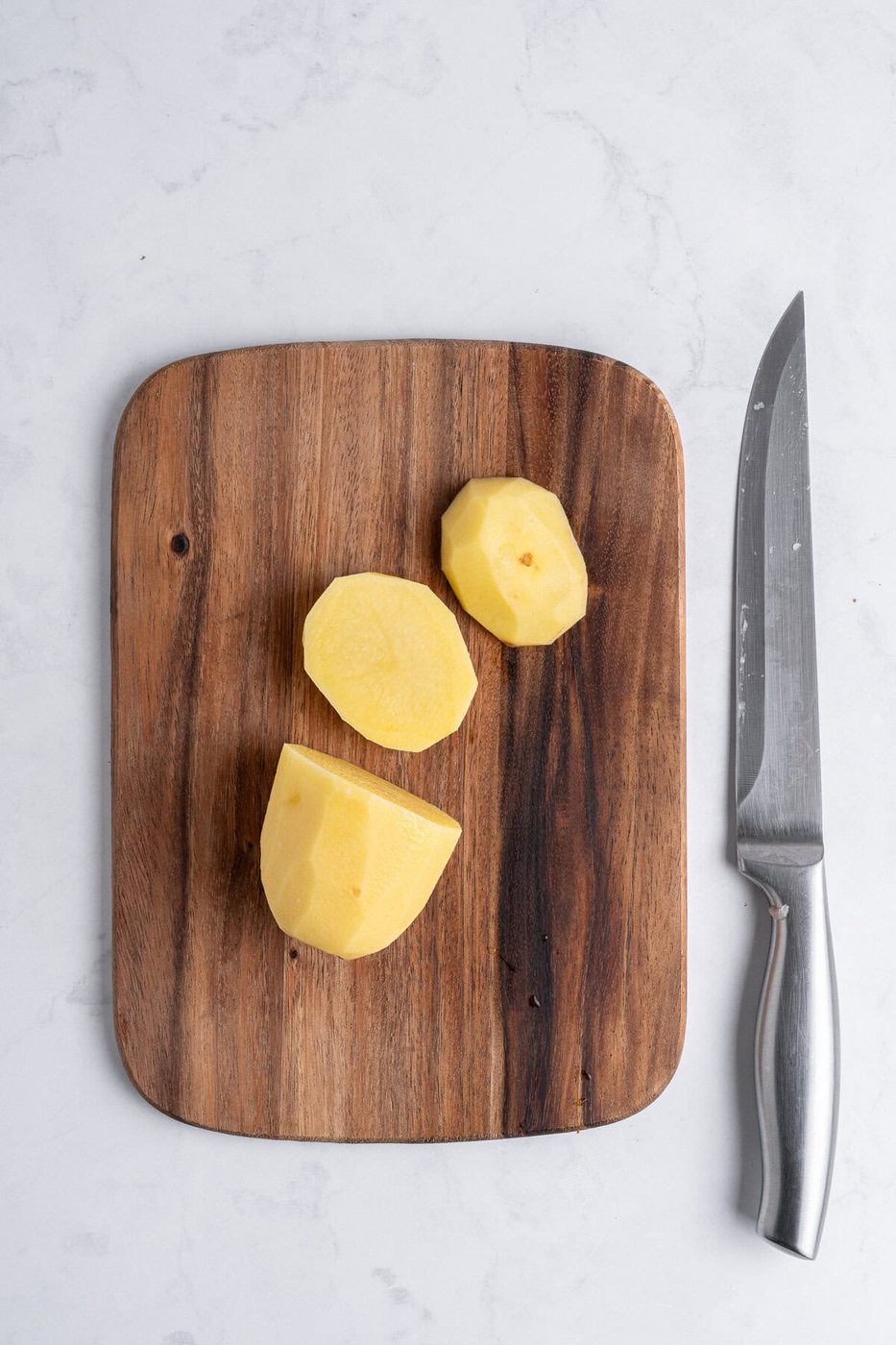 A cutting board with a peeled, cut yellow potato on it and a large chef's knife laying beside it. 