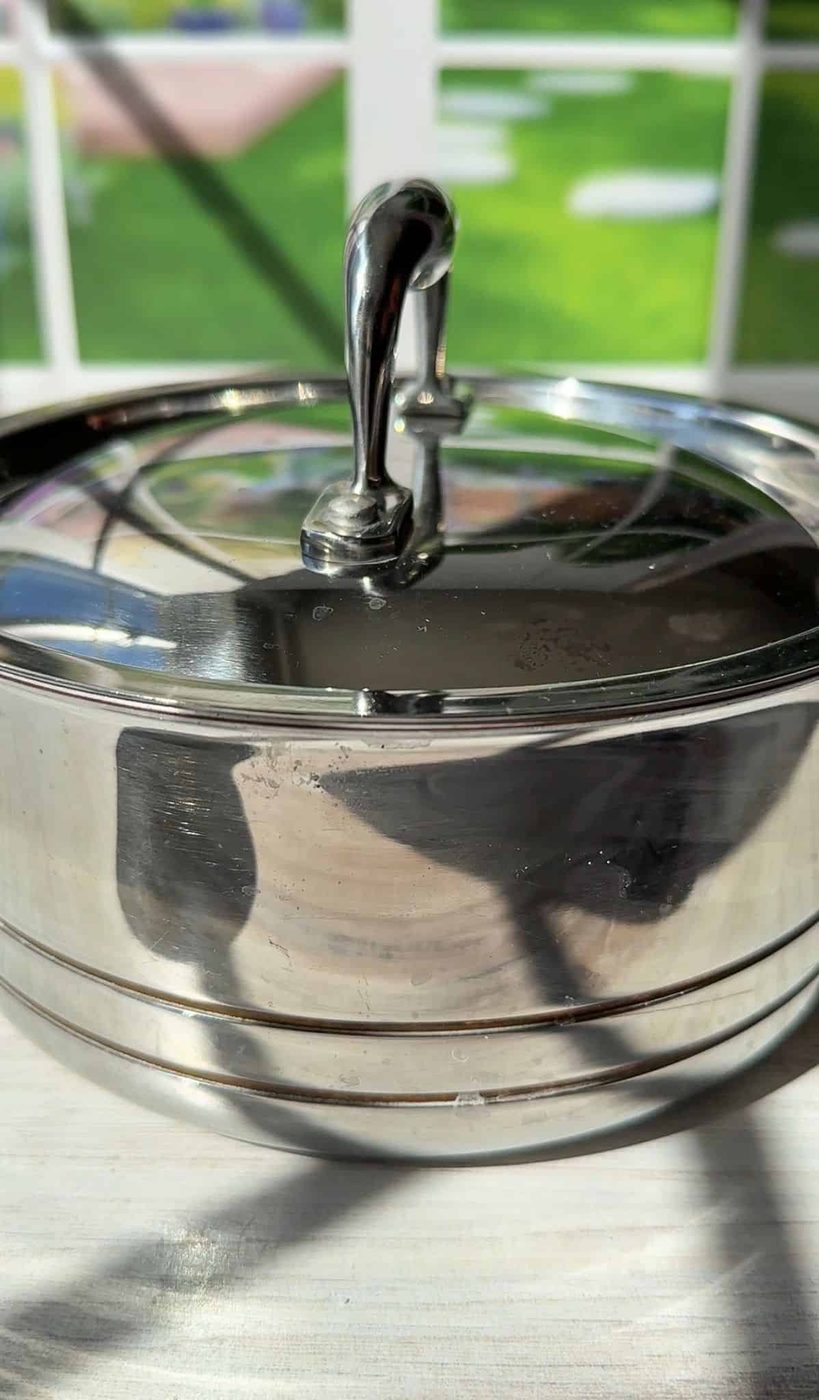 A shiny stainless steel pot with a lid sits on a light-colored surface. The background shows a green lawn and part of a window, with some sunlight casting reflections on the pot's surface. The pot handle is visible, upright on the lid.