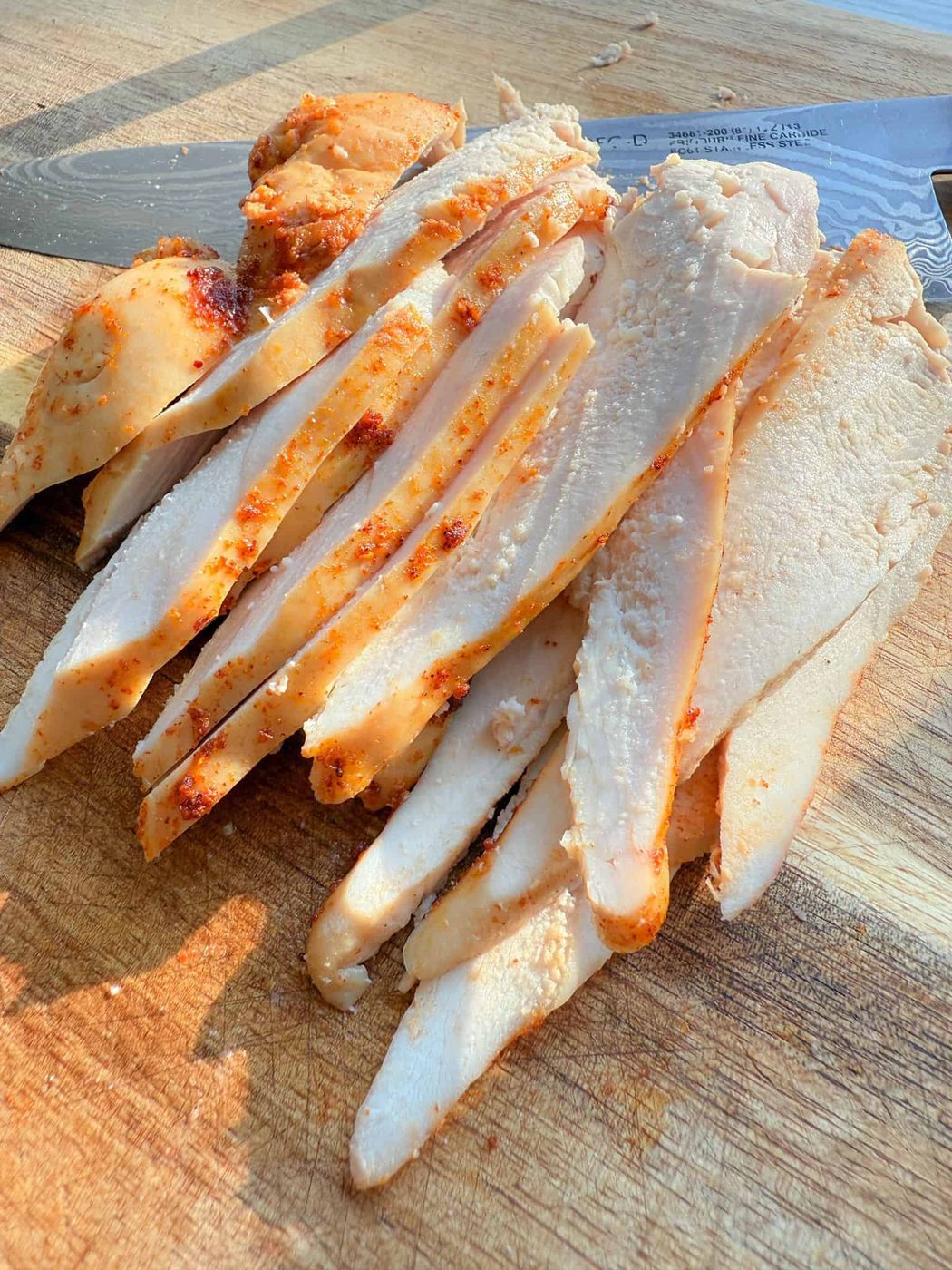 Sliced Chicken Deli Meat arranged on a wooden cutting board with a knife in the background. The chicken appears to be seasoned and cooked, showing a slightly golden-brown exterior and juicy interior. The board has a textured surface with visible grain patterns.
