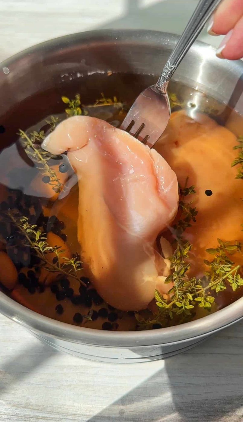 A person uses a fork to lift a raw piece of chicken breast from a bowl filled with water, herbs, and peppercorns. The scene is brightly lit, highlighting the ingredients in the bowl for this Chicken Deli Meat.