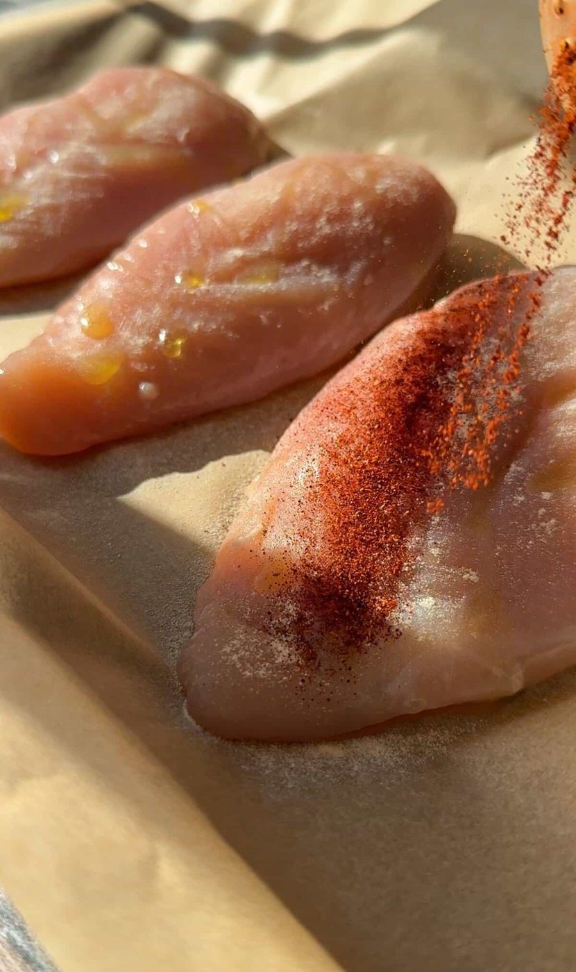 Chicken breasts are arranged in a row on parchment paper. One of the chicken breasts is being sprinkled with a reddish-brown spice, likely paprika, from the right side. The scene is well-lit, highlighting the raw, seasoned chicken.