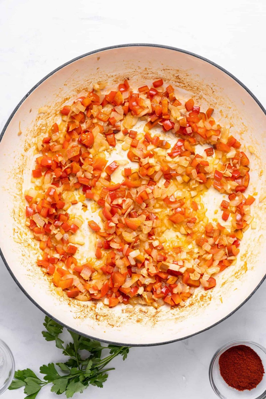 Diced red peppers and onions sautéed in a large pan on a white surface. Fresh parsley and a small bowl of paprika are nearby.