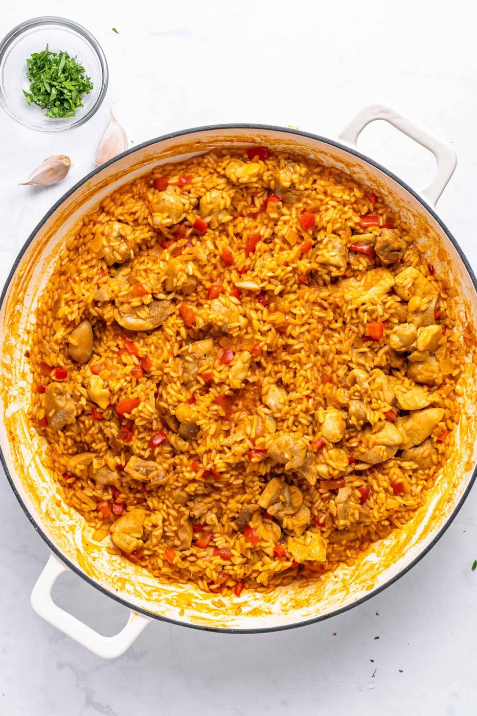 A large pot of vibrant yellow rice mixed with chunks of chicken and red bell peppers, garnished with fresh herbs. It is set on a white marble surface with scattered garlic cloves and a small bowl of chopped herbs nearby.