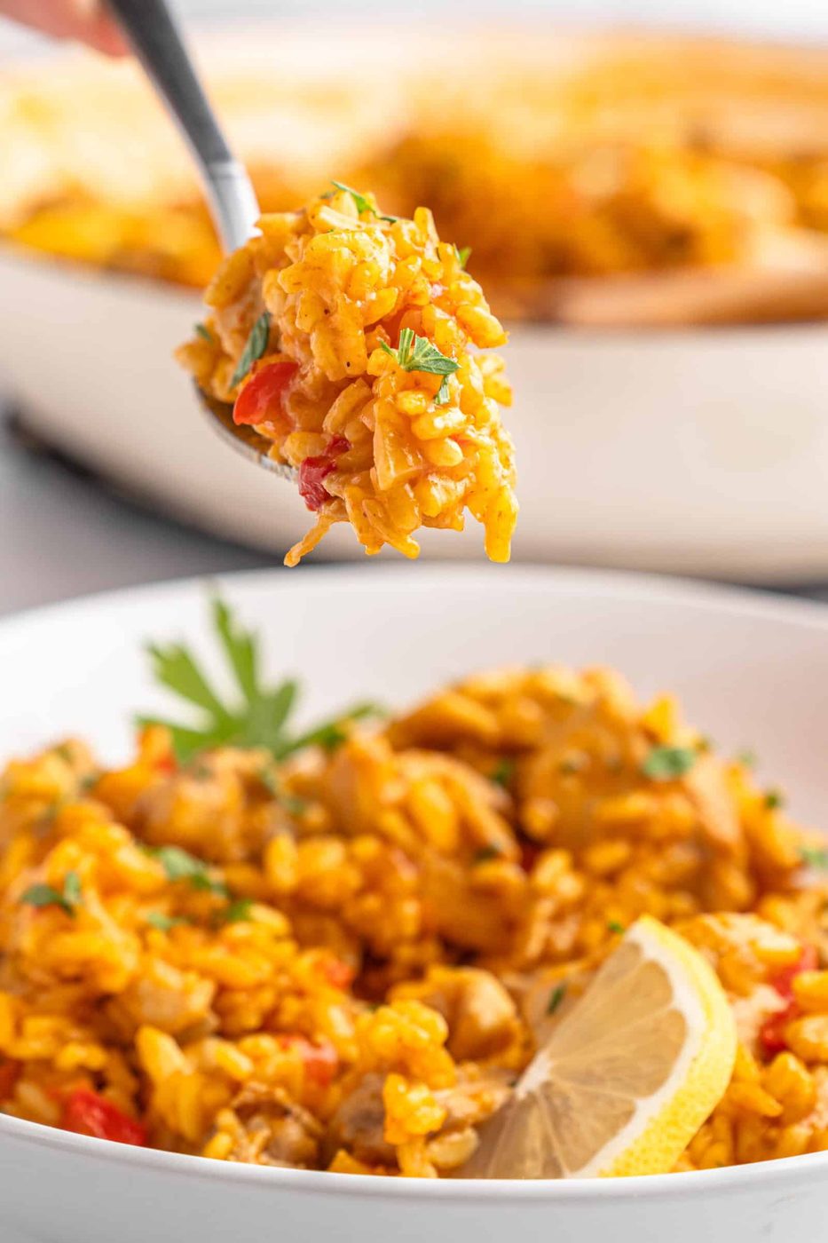 A close-up of a spoon holding a portion of rice over a bowl filled with the dish. The plate features yellow rice, diced vegetables, and garnished with parsley and a lemon wedge. 