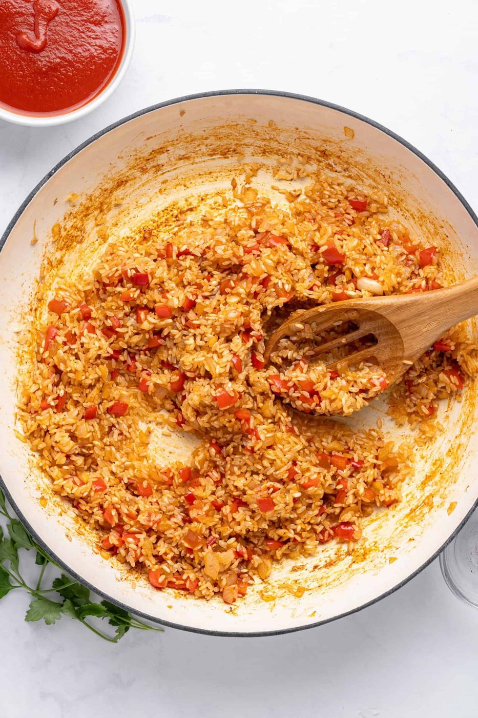 A pot of seasoned rice mixed with chopped red peppers and onions. A wooden spoon rests in the rice. A bowl of tomato sauce is visible in the top left corner. Fresh parsley is partially visible at the bottom left.