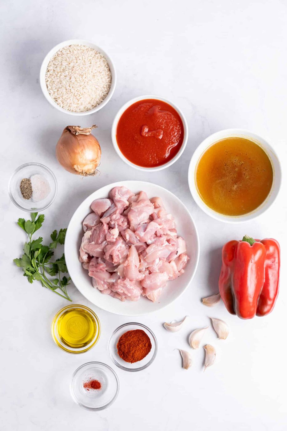 Top-down view of ingredients for a dish: raw chicken pieces in a bowl, white rice, chopped tomatoes, red bell pepper, onion, garlic cloves, chicken broth, oil, spices, and fresh herbs, all arranged on a light surface.