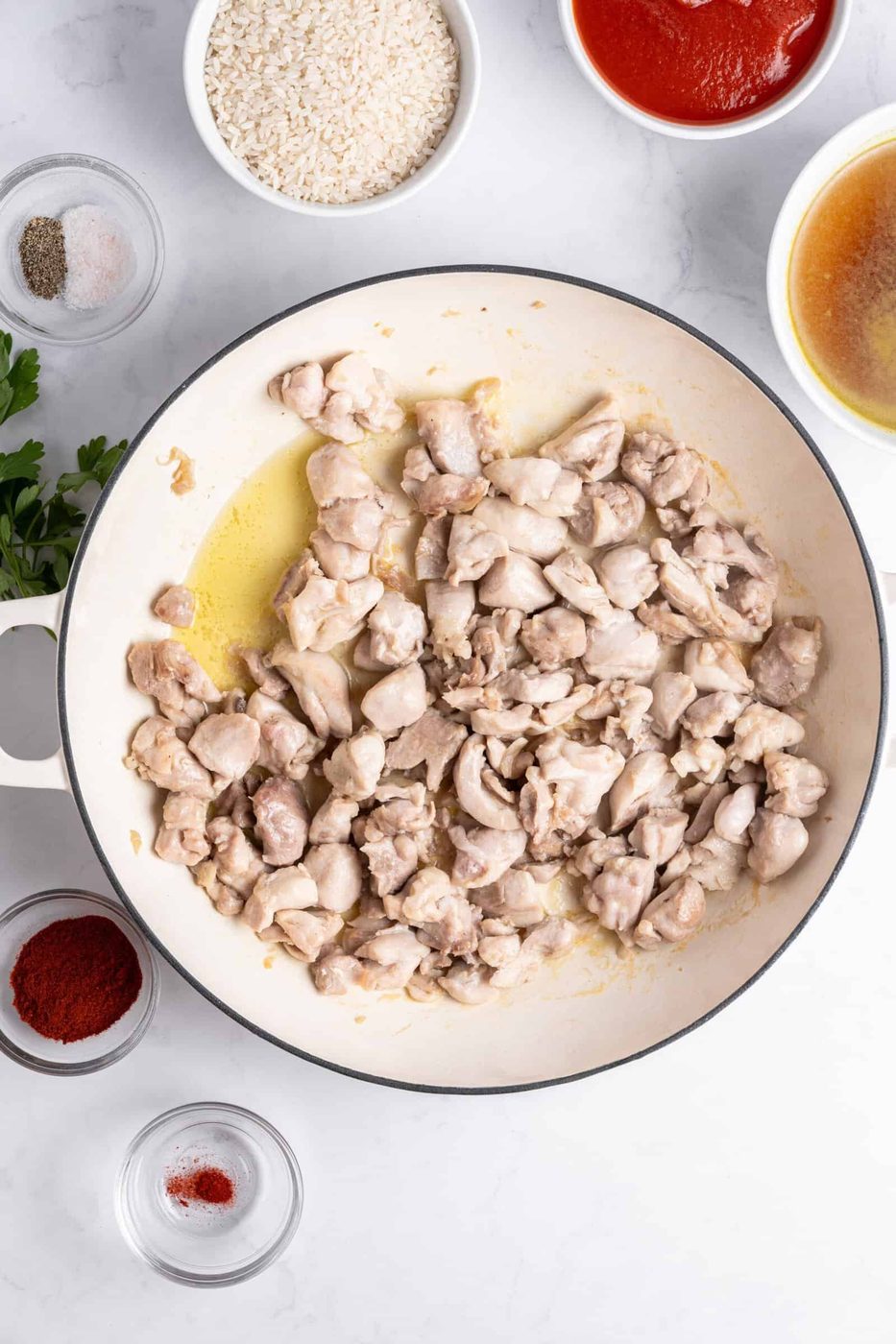 A pan of sautéed chicken pieces on a white surface, surrounded by bowls containing rice, tomato sauce, broth, spices, and fresh parsley.