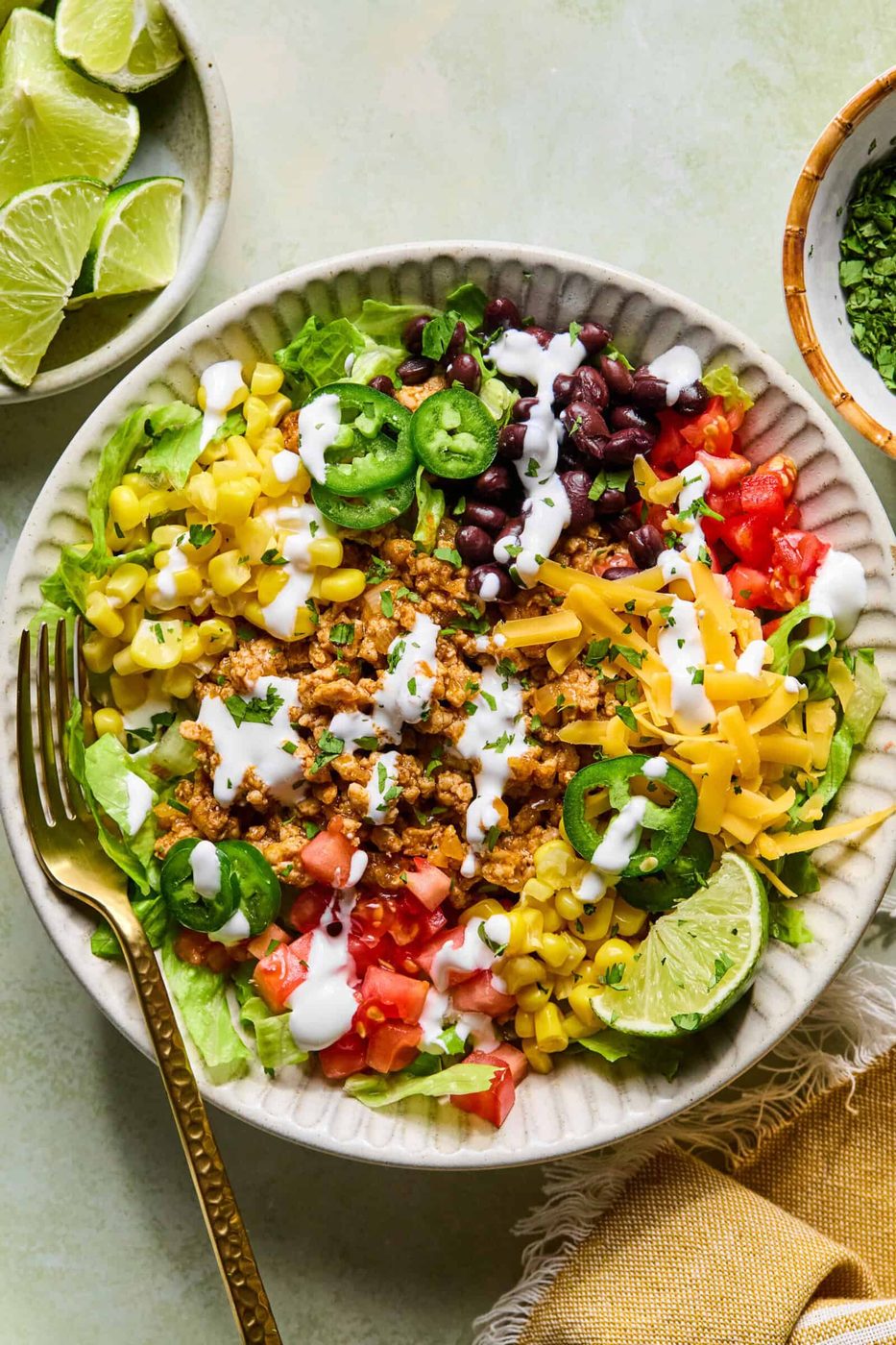 A taco salad in a bowl with ground meat, shredded cheese, black beans, corn, lettuce, tomatoes, jalapeños, and a drizzle of white dressing, garnished with lime wedges. A fork, napkin, and a bowl of lime slices are nearby.