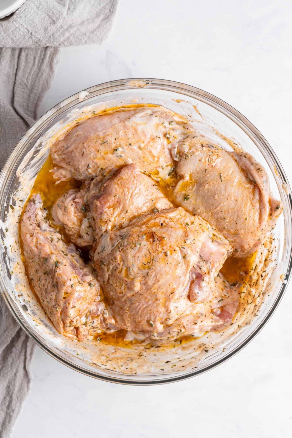 A glass bowl contains raw marinated meat. The meat is coated in a mixture of spices and herbs, giving it a seasoned appearance. The marinating sauce is visible at the bottom of the bowl, and a light cloth is partially visible beside it.