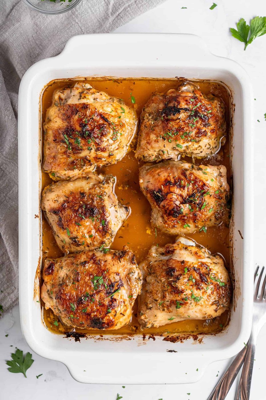 Baked chicken thighs in a white rectangular dish, topped with herbs and surrounded by a golden sauce. A gray cloth and silverware are placed beside the dish on a marble countertop.