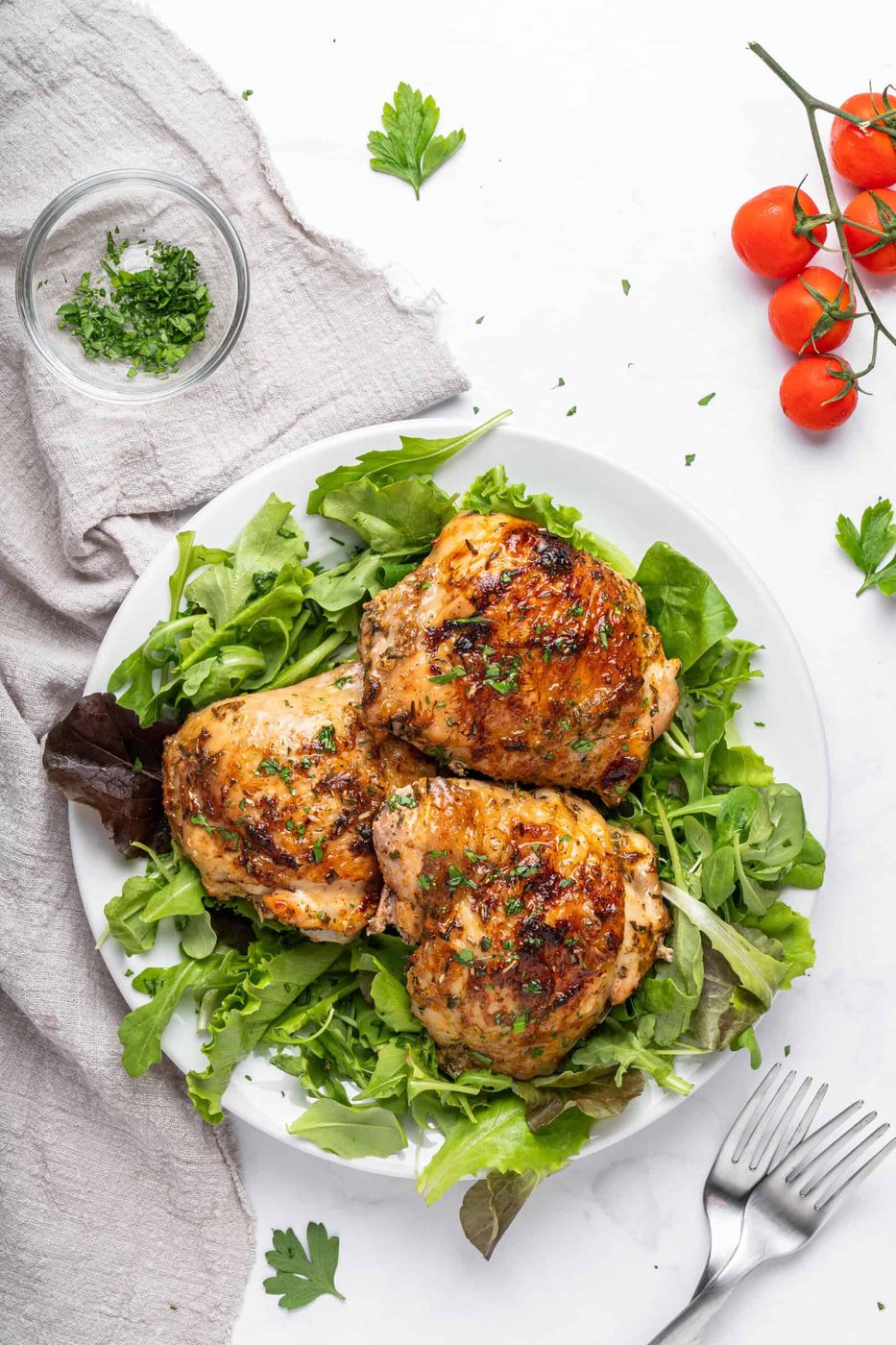 A plate with three grilled chicken thighs garnished with herbs sits on a bed of mixed greens. Nearby are cherry tomatoes on the vine, a small bowl of chopped herbs, a fork, and a gray napkin on a white marble surface.