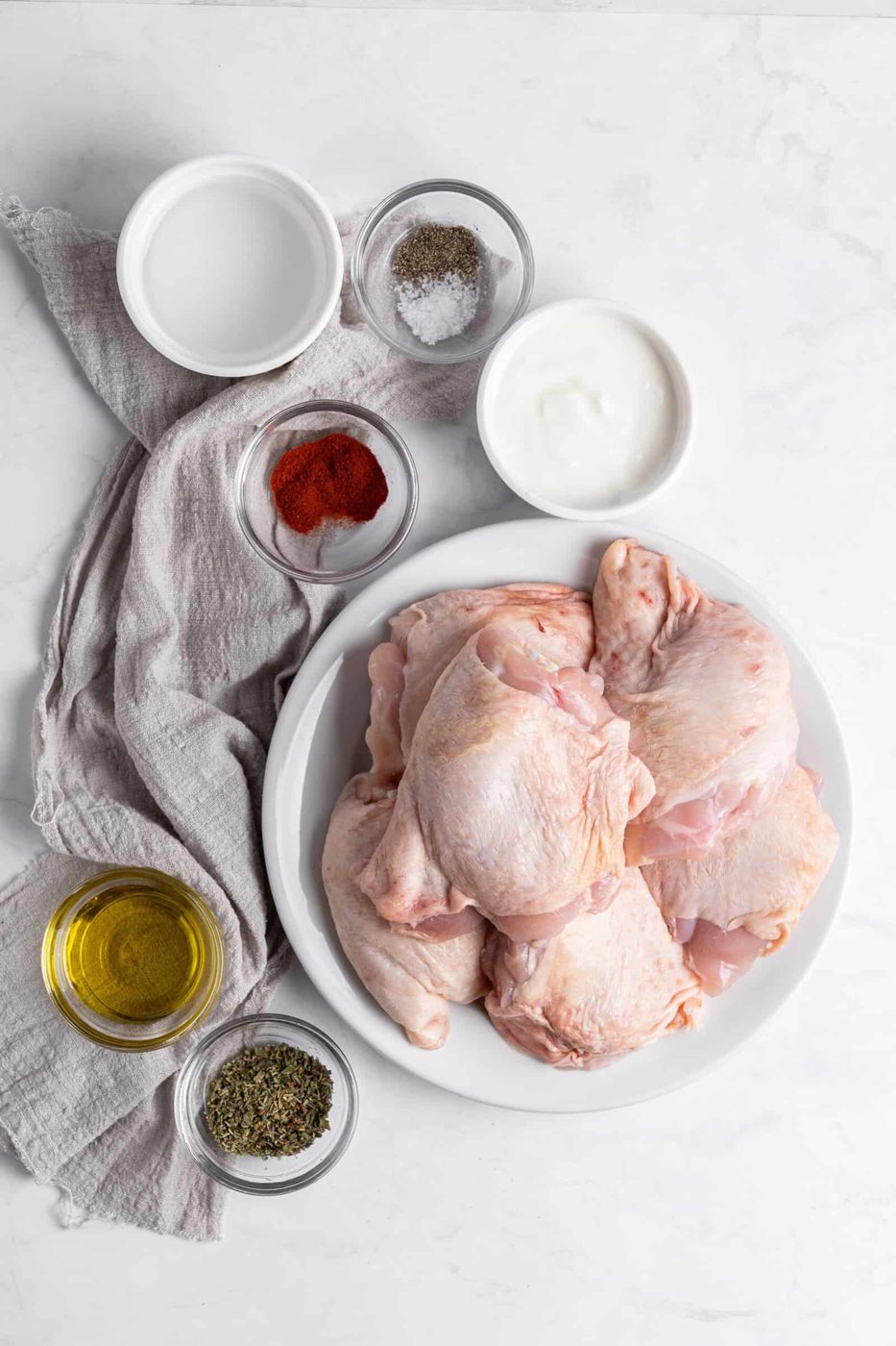 Raw chicken thighs on a plate surrounded by small bowls of olive oil, yogurt, salt, pepper, paprika, and herbs on a marble surface. A gray cloth napkin adds texture to the setup.