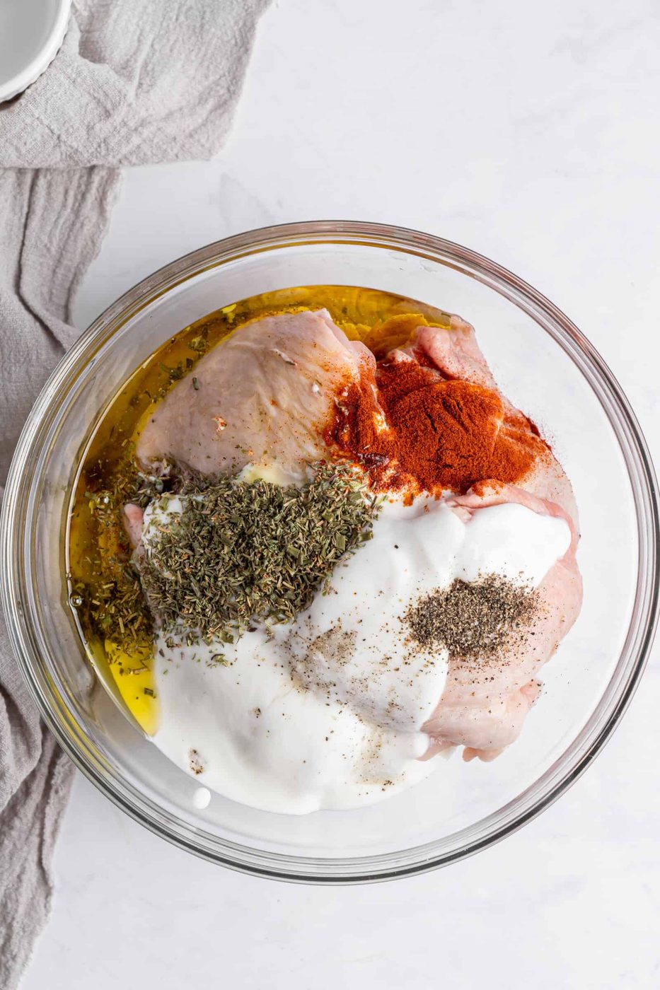 A glass bowl contains raw meat, yogurt, olive oil, paprika, mixed herbs, and black pepper. A light gray fabric is partially visible on the left, set on a white marble surface.