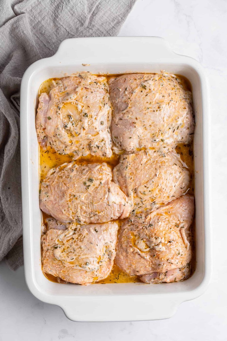 Marinated meat in a white rectangular baking dish on a marble surface, with a gray cloth nearby. The marinade includes herbs and oil.