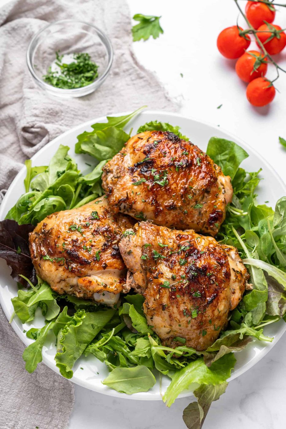 Grilled chicken thighs garnished with herbs are served on a bed of mixed greens on a white plate. A small bowl of chopped herbs and a bunch of fresh cherry tomatoes are in the background.