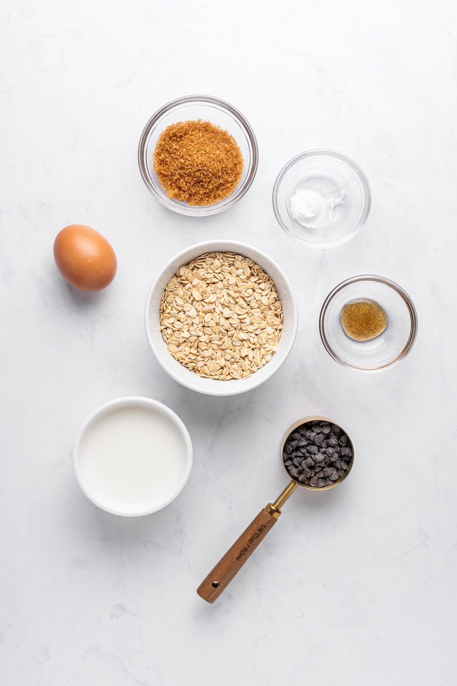 Ingredients for baking arranged on a white surface: an egg, a bowl of rolled oats, brown sugar, a small bowl of baking powder, a pinch of ground spice, a cup of milk, and a scoop of chocolate chips.