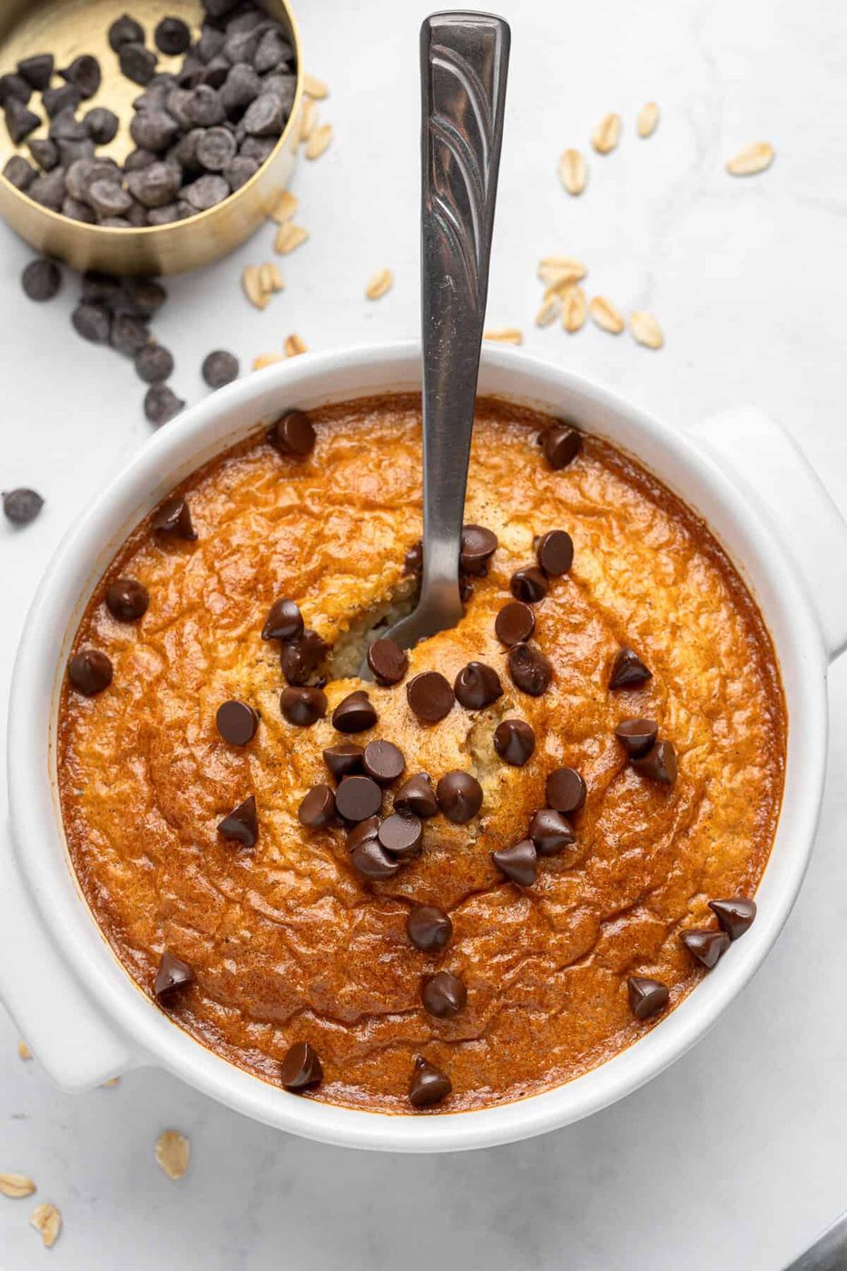 A bowl of baked oats topped with melted chocolate chips. A spoon is dipped into the oats, and there are chocolate chips scattered on top. A small bowl of extra chocolate chips is visible in the background on a light marble surface.