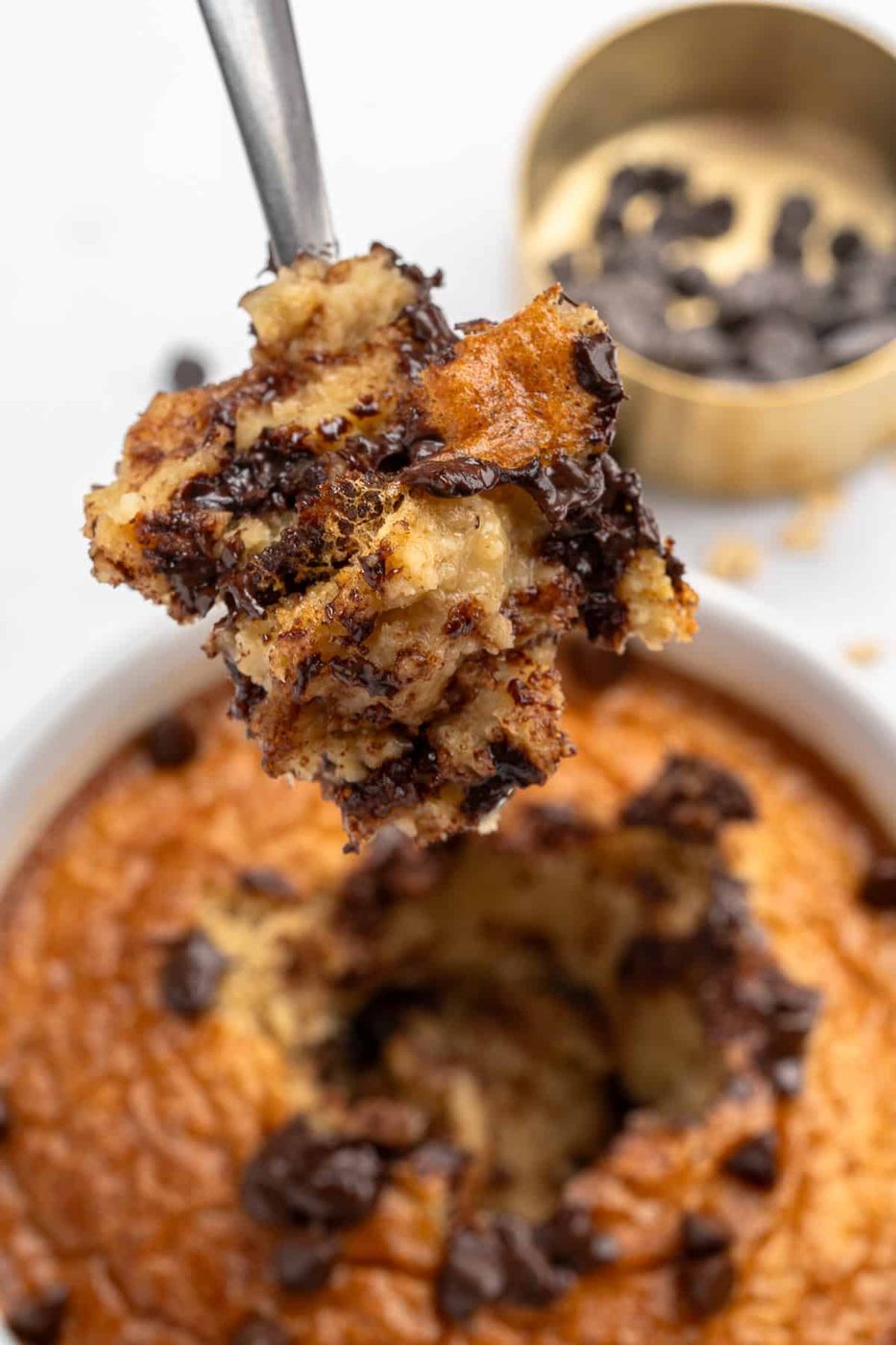 A spoon holds a bite of chocolate chip banana bread pudding above a bowl filled with the dessert. In the background, a small container of chocolate chips is visible. The bread pudding is golden brown with scattered chocolate chips.