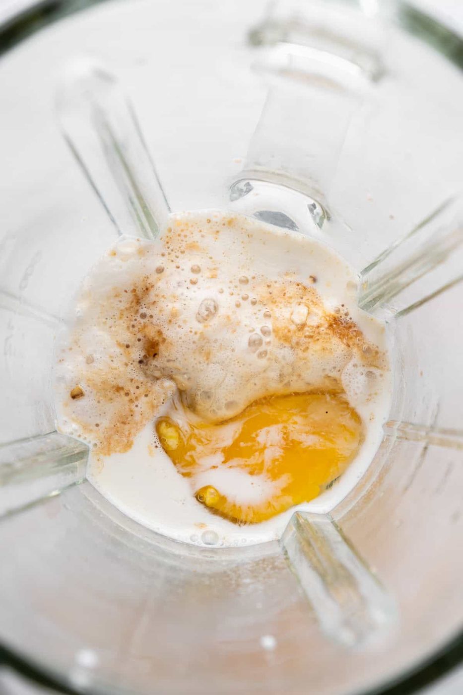 Top view of a blender with egg, milk, and spices inside. The ingredients are partially mixed, creating a frothy texture. The blender's glass walls are visible, showcasing the creamy liquid and spices.