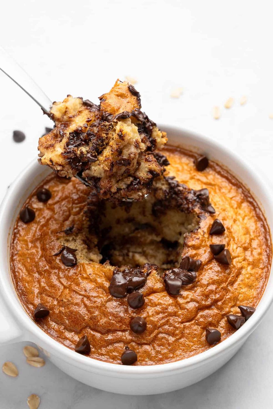 A close-up of a baked dessert in a white bowl. The oatmeal is golden-brown. A spoon lifts a portion, revealing the moist, chocolatey interior.