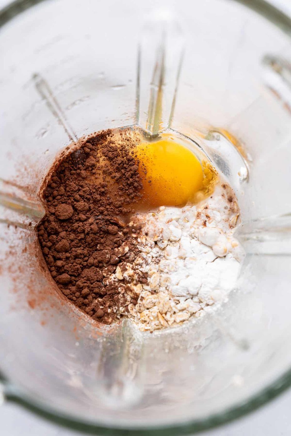 Top view of a blender containing ingredients for baking, including cocoa powder, an egg, flour, oats, and a liquid. The ingredients are placed in different sections, ready to be blended.