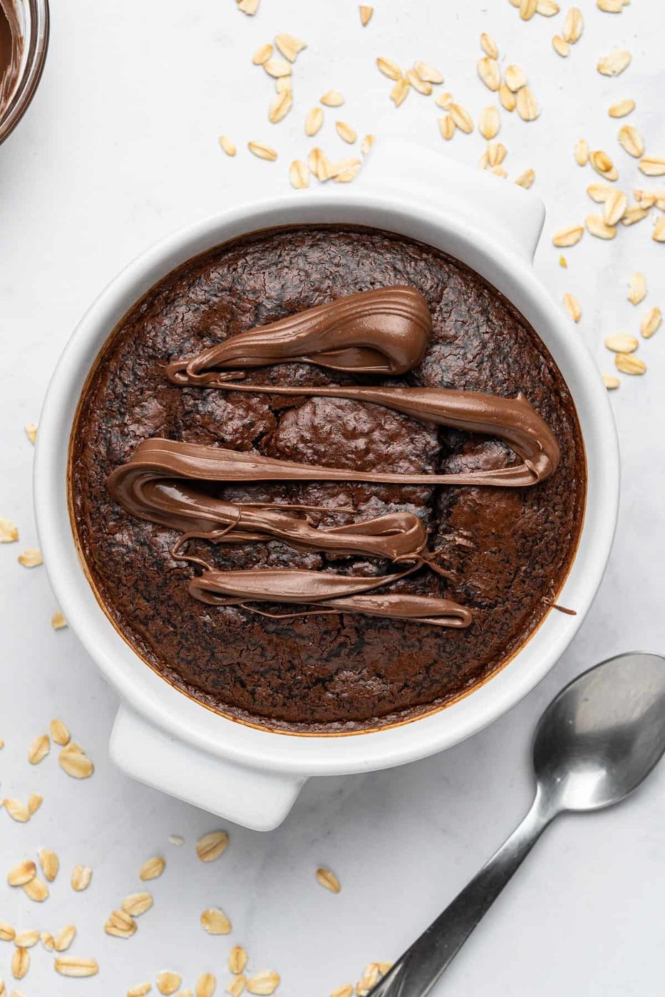 A round white bowl filled with a baked chocolate oat dessert, topped with drizzles of smooth chocolate. A spoon lies beside the bowl on a white surface, scattered with oats.