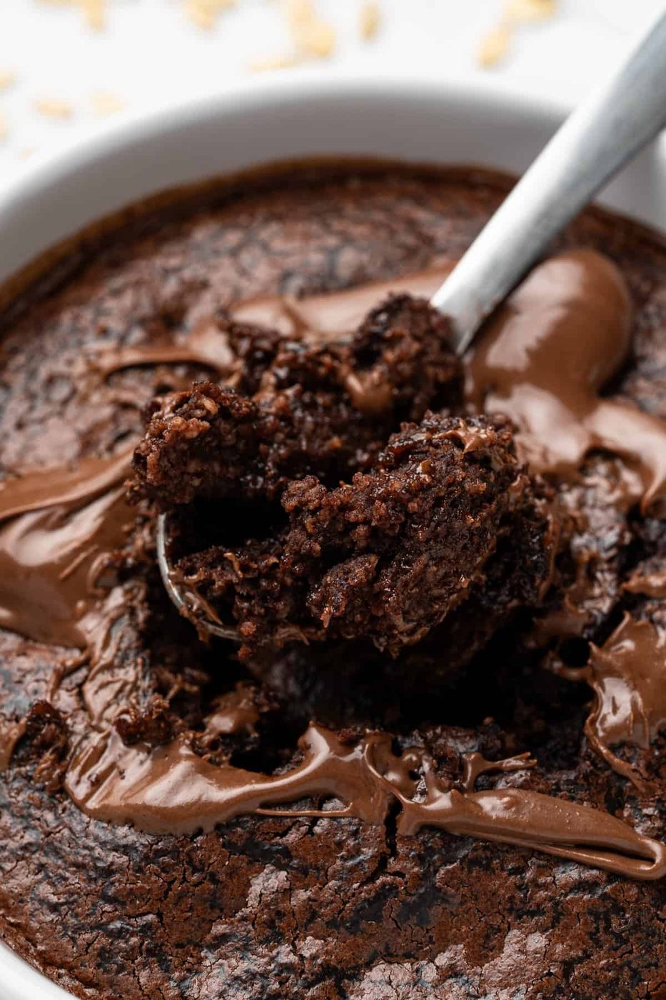 A close-up of a spoon scooping a portion of gooey, baked chocolate dessert from a white dish. The surface is rich and textured, with melted chocolate swirls. The background is blurred with a hint of scattered ingredients.