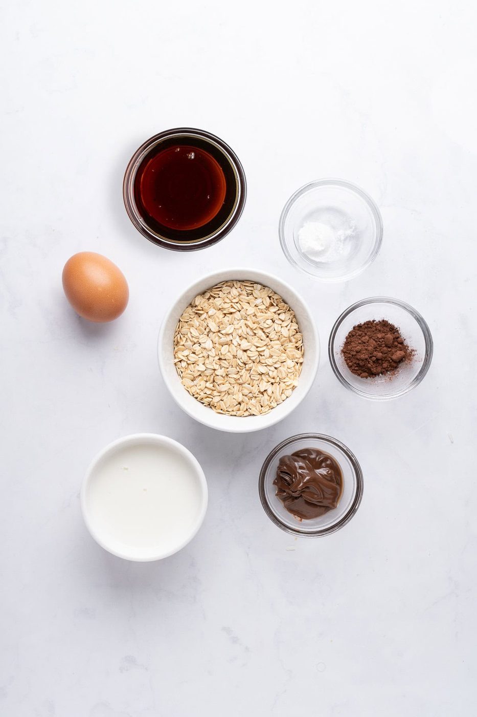 A flat lay of baking ingredients on a white surface. Includes a bowl of oats, a glass of milk, an egg, small bowls of cocoa powder, baking soda, chocolate spread, and a bowl of maple syrup.