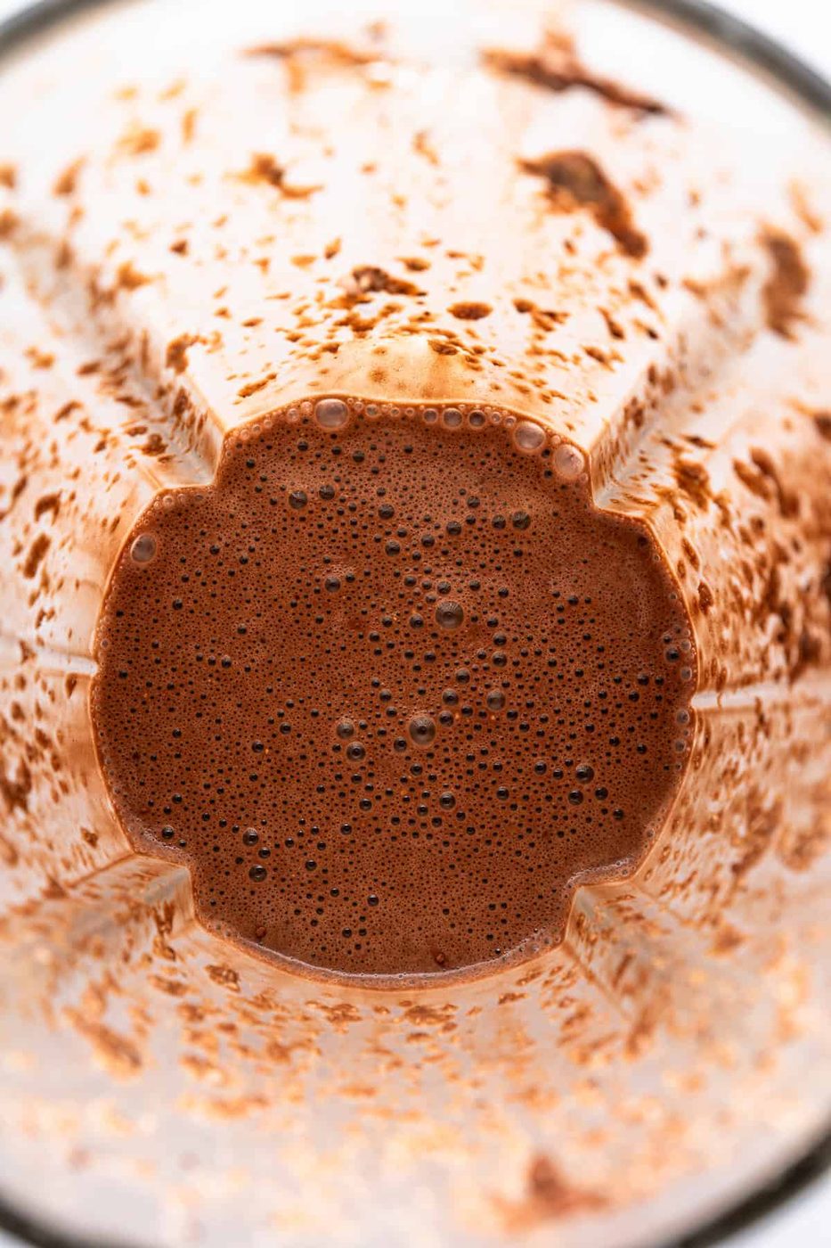 A close-up view of a blender container with chocolate smoothie remnants clinging to the sides. The bottom shows a frothy, rich chocolate mixture with small bubbles.