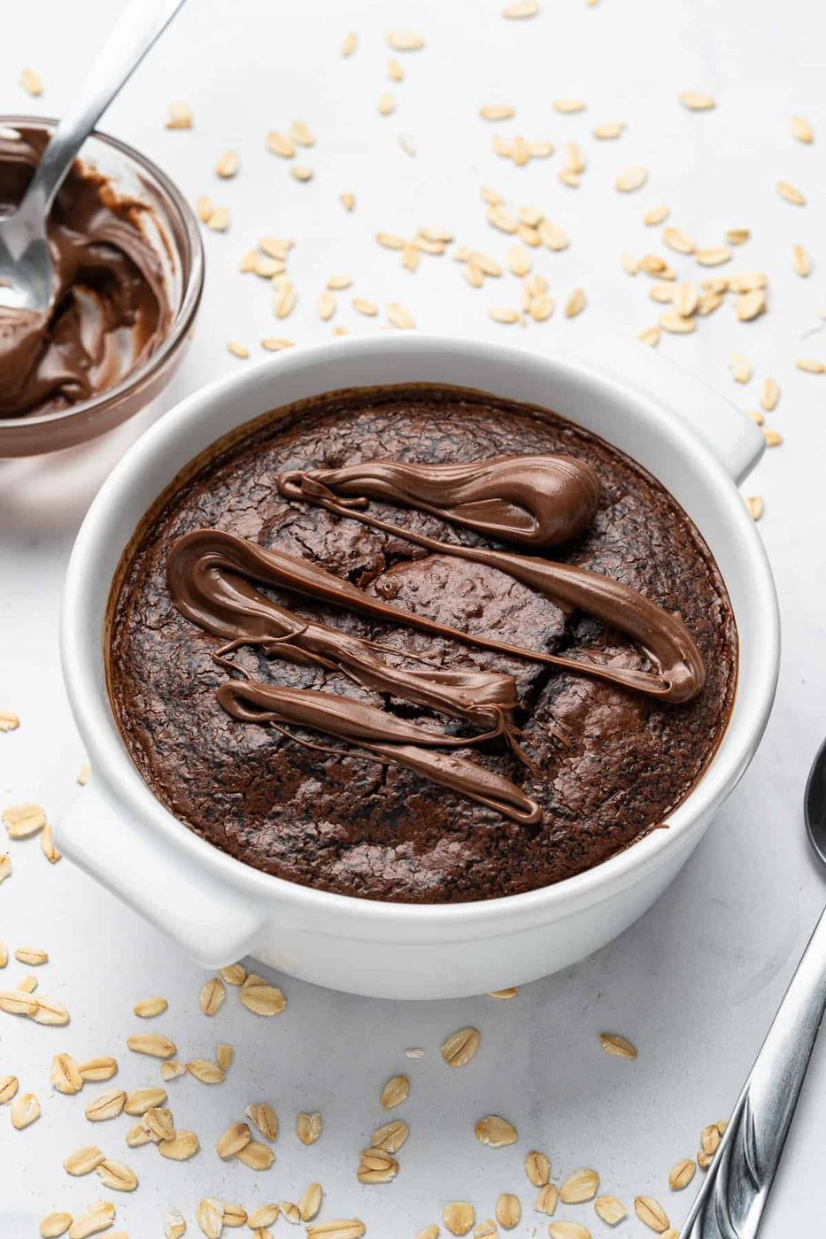 A baked oatmeal dish topped with drizzled chocolate sits in a white bowl. A spoon rests beside it on a white surface scattered with oats. A bowl of chocolate spread is nearby.
