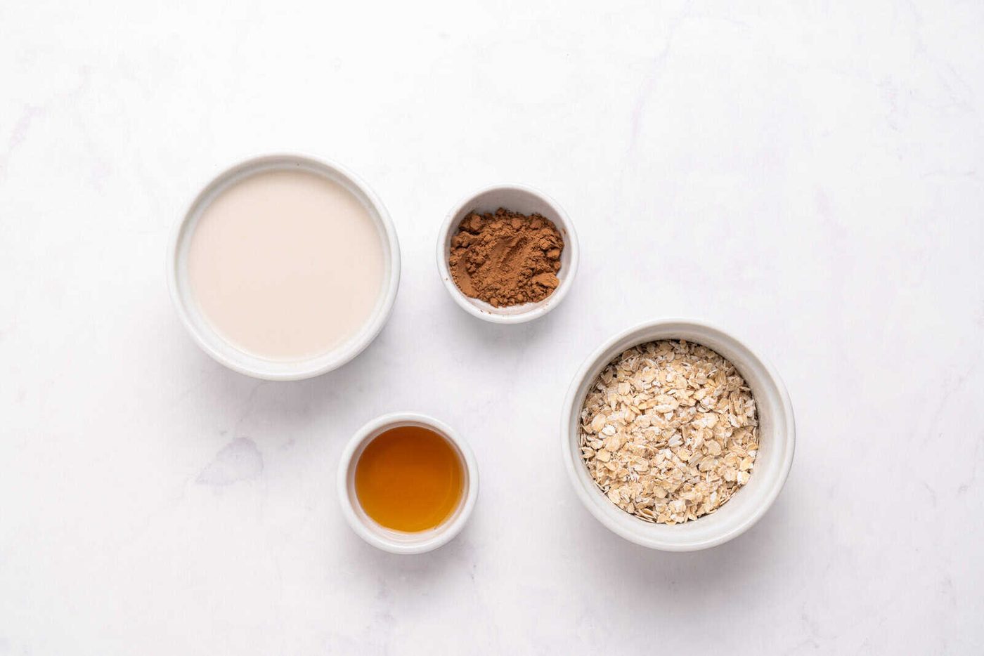 Four small bowls on a white surface containing oat milk, cocoa powder, rolled oats, and maple syrup, arranged in a flat lay style.