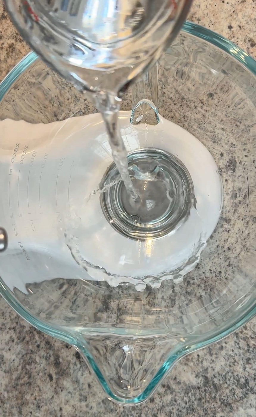 A stream of clear liquid is being poured from a glass into a large glass measuring bowl on a speckled countertop, the first step in preparing ingredients for a Cloud Bread.