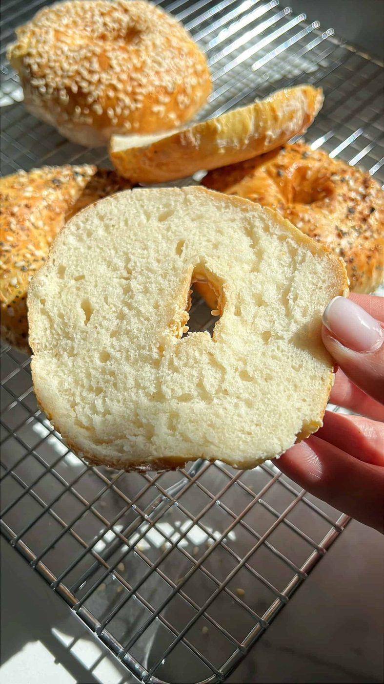 A close-up of a sliced cottage cheese bagel held by a hand. The bagel is on a cooling rack, surrounded by whole bagels with various toppings like sesame seeds and herbs. Sunlight casts shadows, creating a warm ambiance.