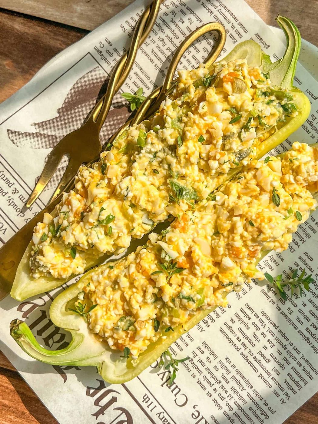 Two halved green peppers stuffed with egg salad are served on a sheet of newspaper print, with a gold fork and knife placed alongside on a wooden surface. Sunlight casts a warm glow over the dish.
