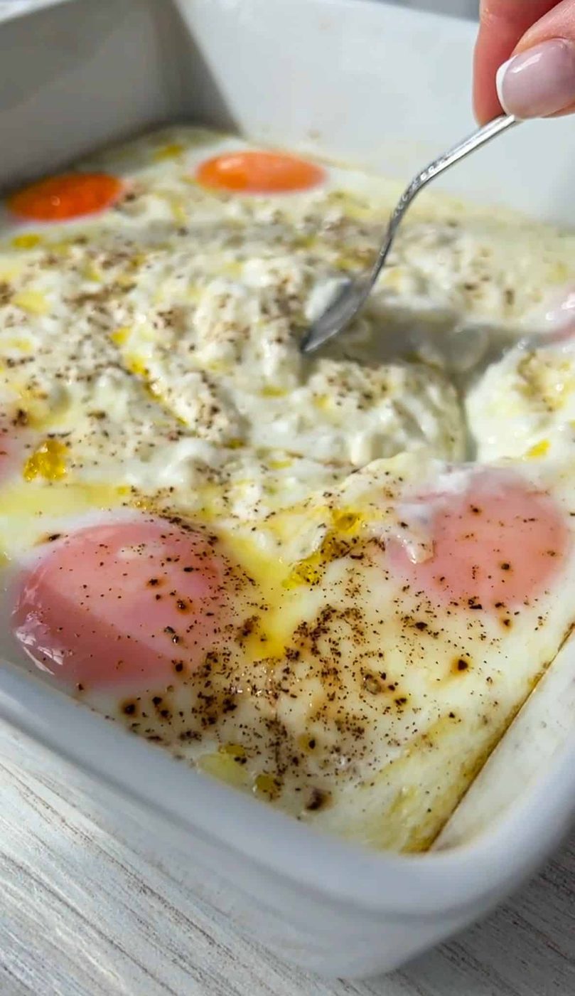 A spoon is stirring a dish with poached eggs seasoned with pepper in a white baking dish. The eggs are nestled in a creamy sauce.