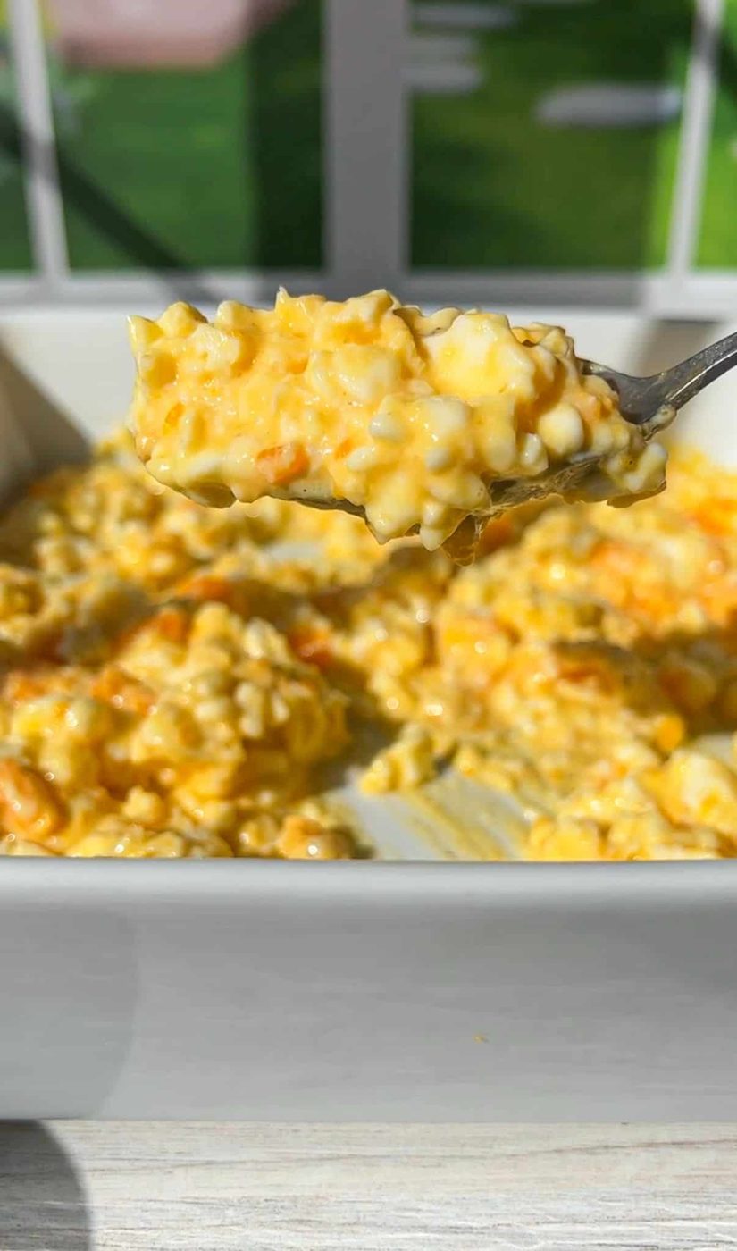 A spoonful of cottage cheese eggs is held above a white baking dish. The dish is filled with creamy, golden casserole, with visible chunks of potatoes and melted cheese. A sunlit window is blurred in the background.