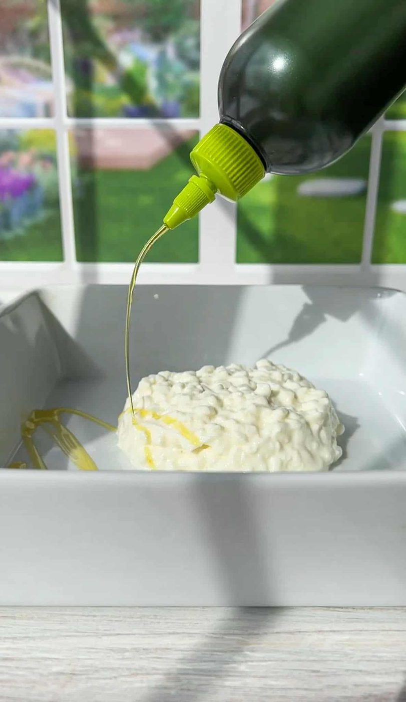 A black bottle with a green nozzle pours oil over a round piece of burrata cheese in a white square dish. The background features blurred garden scenery visible through a window.